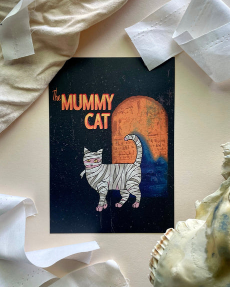 A print of a sphynx cat with a mummy-like wrapping, positioned in front of a skull and some bandage-like paper, suggesting a classic horror theme.