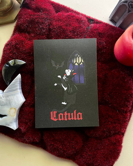 A black and white art print of a cat with a vampire-themed backdrop, titled 'Catula', placed on a red textured surface with a candle and a skull decoration in the background.