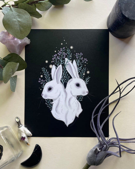 An art print of a two-headed rabbit with stars and a branch in the background, placed on a surface with a bottle, plant, and crystal.