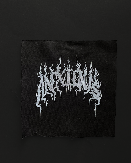 A black patch with the word 'ANXIOUS' written in a stylized font, hand screen printed design, with unfinished edges.