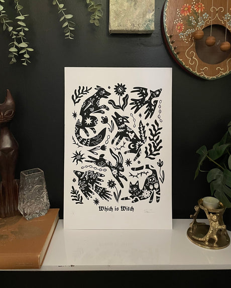 Black and white art print titled 'Which is Witch' on a white stand against a dark wall with decorative items.