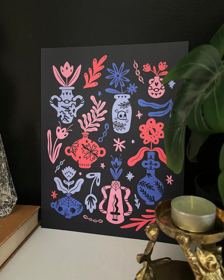 Decorative print with colorful vases and floral patterns on a black background, placed on a surface with a plant and decorative items.