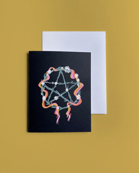 A greeting card with a black background featuring a colorful pentagram wreath design and 'Beltane' text, accompanied by a white envelope.