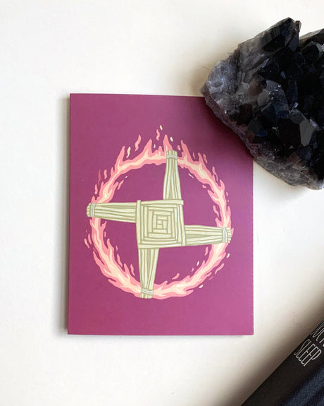 A card with a hand-drawn illustration of Brigid's Cross on a purple background, accompanied by a black crystal and a dark book on a white surface.