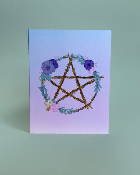 A greeting card with a hand-drawn illustration of a wooden pentacle surrounded by flowers and foliage on a purple background, with a plain white envelope.