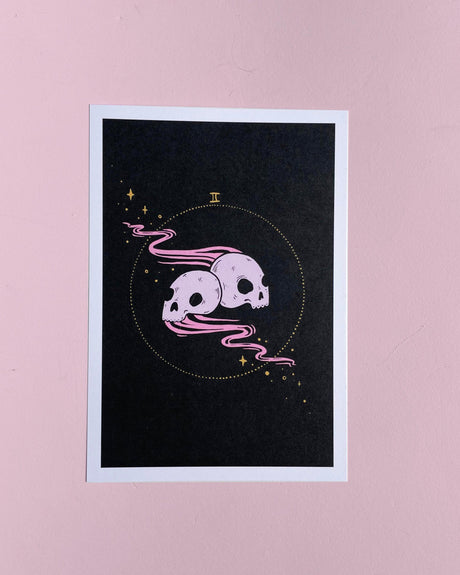 A black card with a hand-drawn illustration of two back-to-back skulls connected by a pink line, representing the Gemini zodiac sign, placed against a pink background.